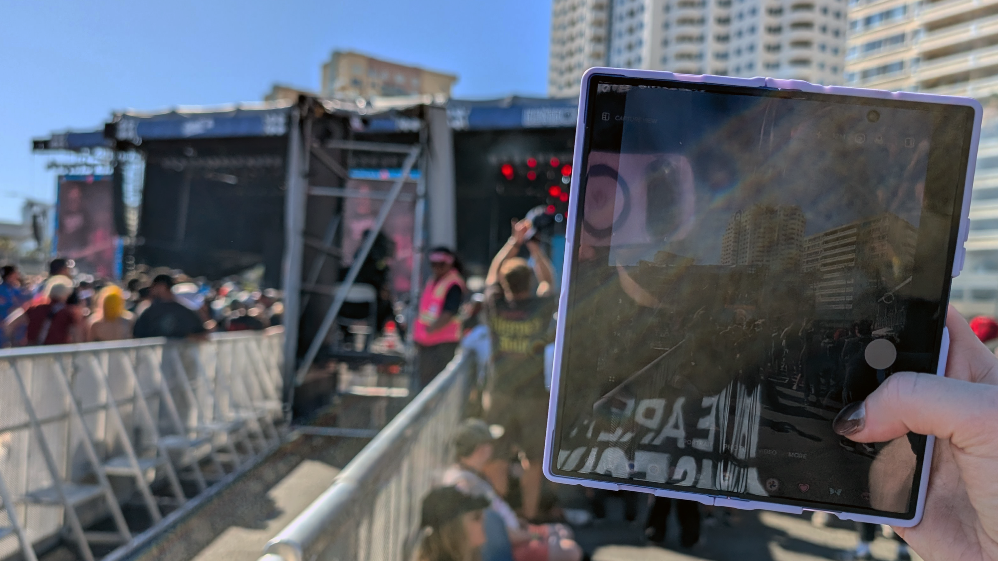 A photo of Auntie holding up a tablet-like device to snap a photo of some dudes on a stage