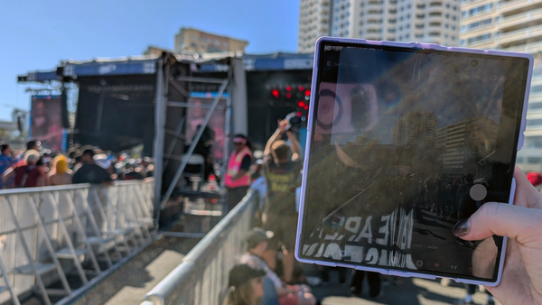A photo of Auntie holding up a tablet-like device to snap a photo of some dudes on a stage